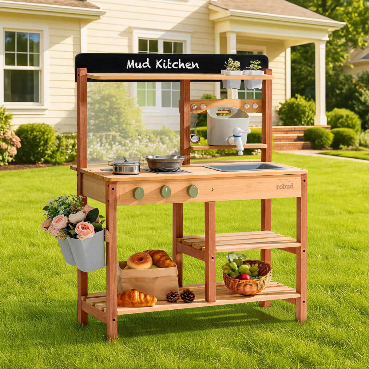 ROBUD outdoor mud kitchen standing on grass in front of a house, featuring a chalkboard header, sink area, hanging pots, and flower bucket on the side