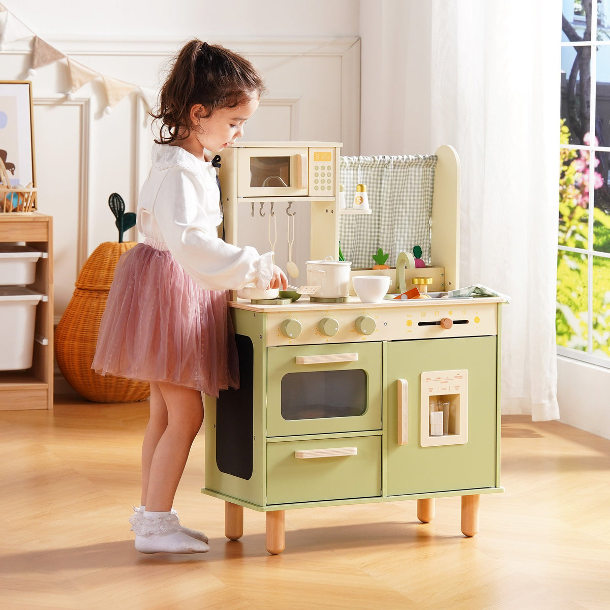 Girl in a pink tutu stands at the ROBUD sage green wooden play kitchen and opens the refrigerator door compartment.