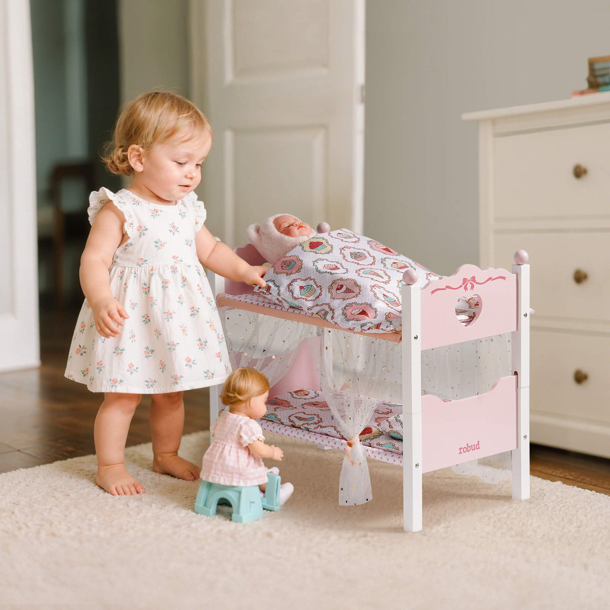 Little girl in a floral dress stands beside the ROBUD pink princess doll bunk bed placing a baby doll on the top bunk tier