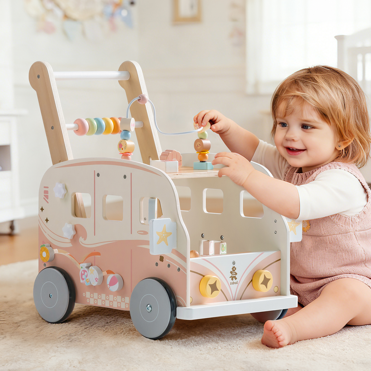 Seated toddler plays with bead and block activities on the ROBUD pink bus walker toy in a cosy home setting.