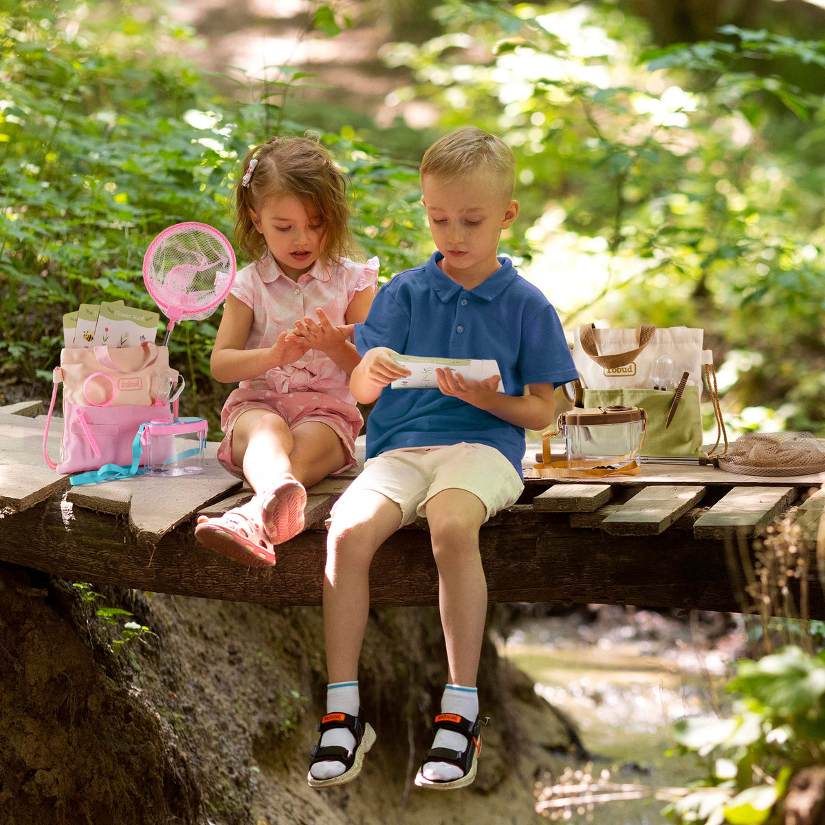 Girl and boy in hats crouching in sunlit forest using ROBUD pink magnifying glass to study a ladybug on a leaf together