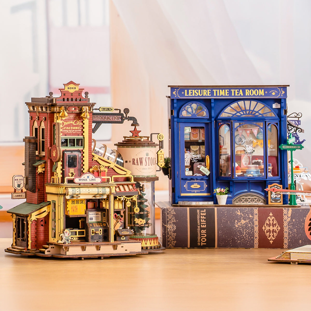Rolife miniature house lifestyle scene with two illuminated DIY kits on display including a red Victorian-style factory and a blue Leisure Time Tea Room shop