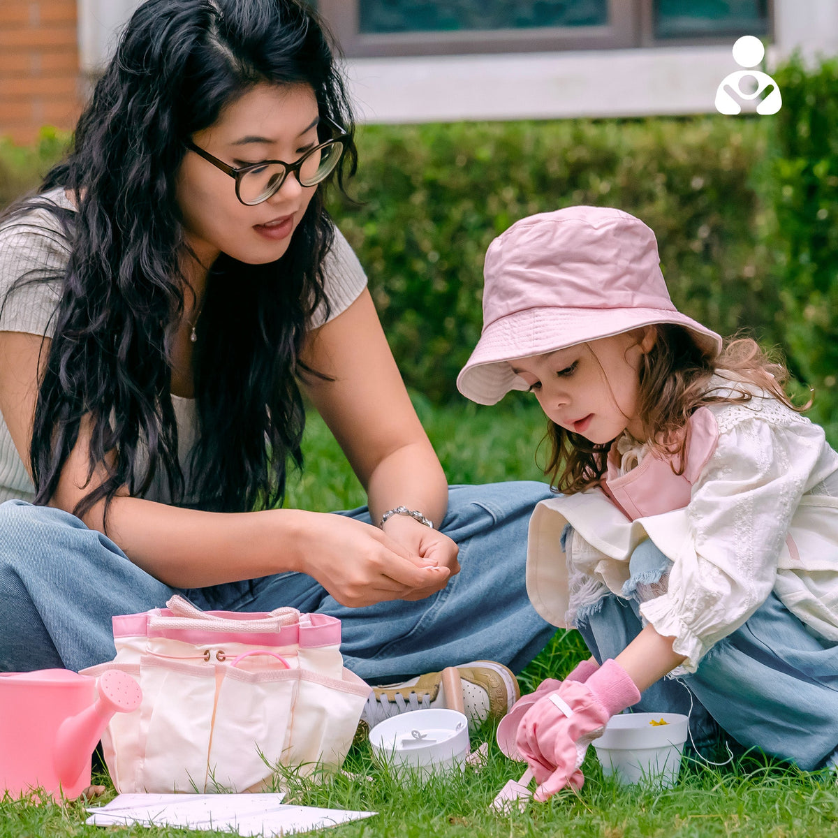 Girl in pink bucket hat kneeling on grass digging soil with ROBUD Kids Gardening Set pink shovel into a white potting cup