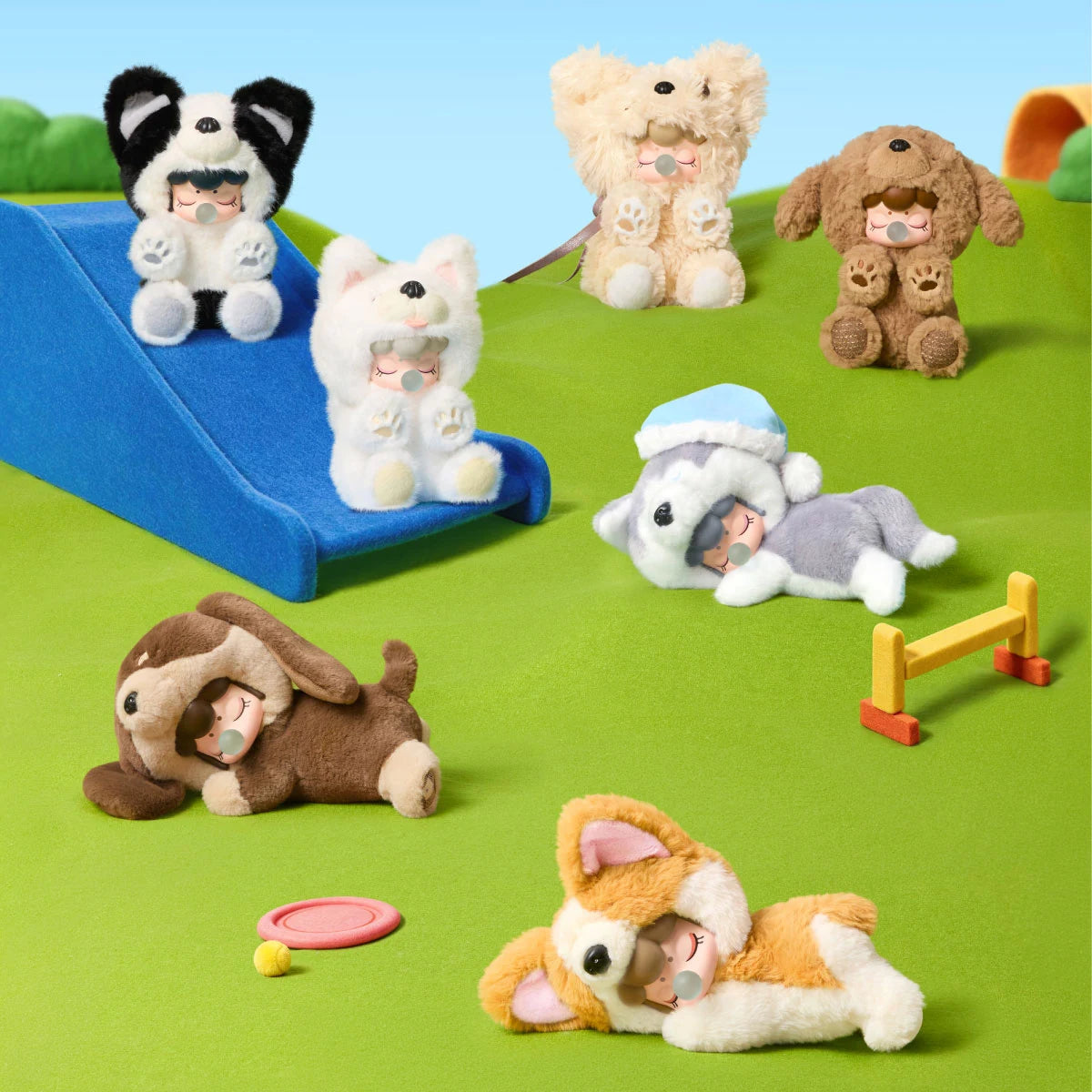 Sechs Plüschfiguren in Hundekostümen mit geschlossenen Augen und Blasen-Accessoires posieren auf einem grünen Filzspielplatz mit blauer Rutsche, Wippe und Ball.