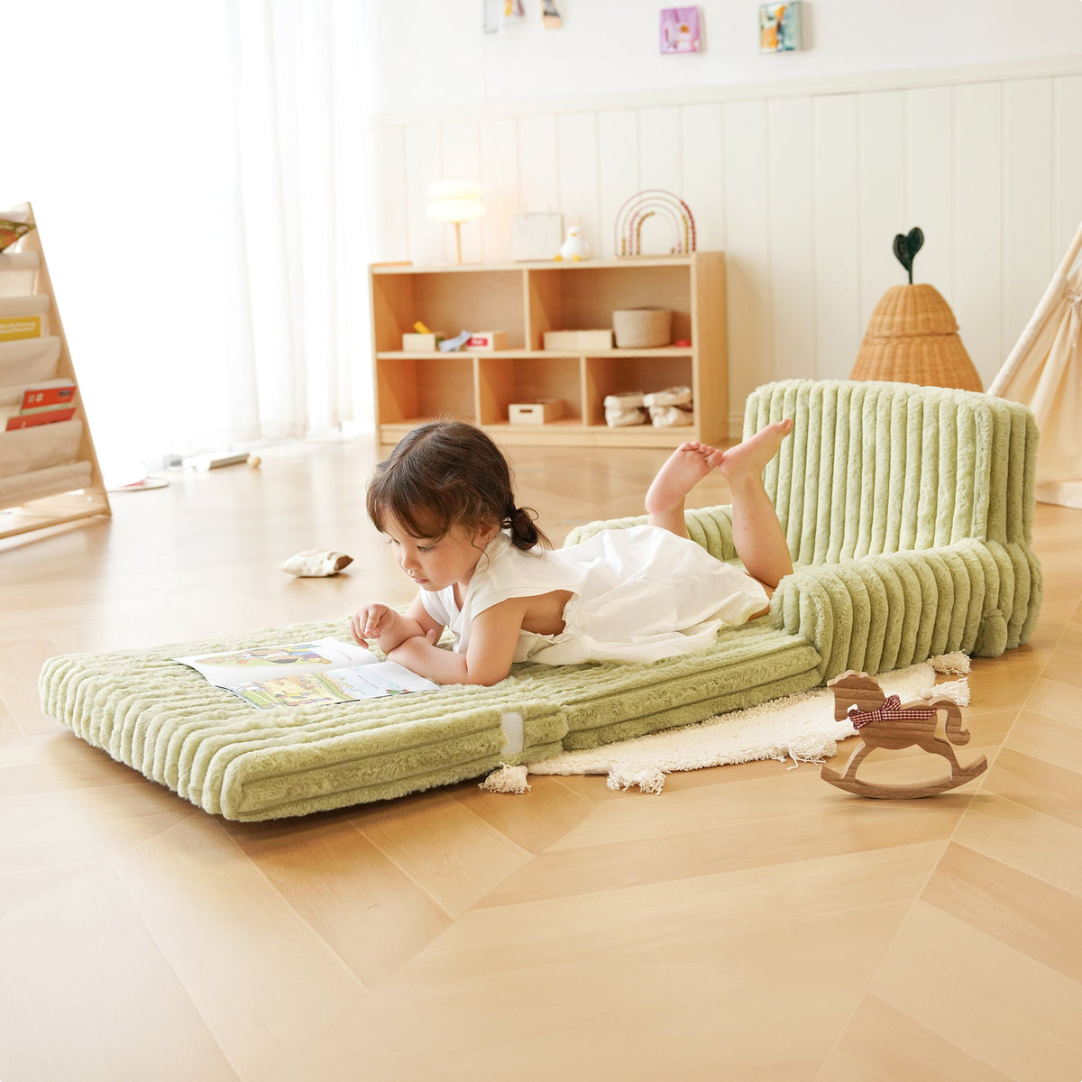 Young girl in a white dress lying on an unfolded ROBUD green striped sofa mattress reading in a bright playroom.