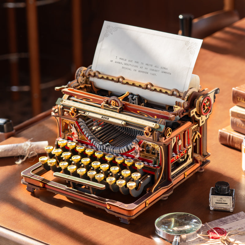 Robotime wooden typewriter model kit with rolled paper in the carriage, on a desk beside books and a magnifying glass