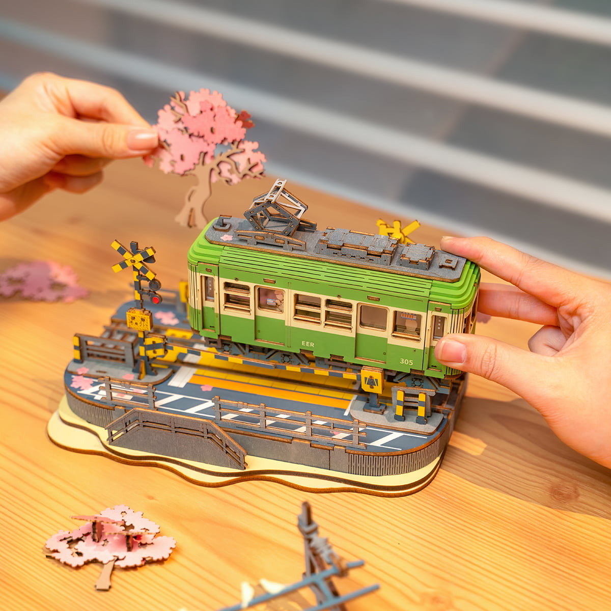 Two hands assembling the Rolife Sakura Journey tram diorama on a wooden table with scattered pink cherry blossom pieces