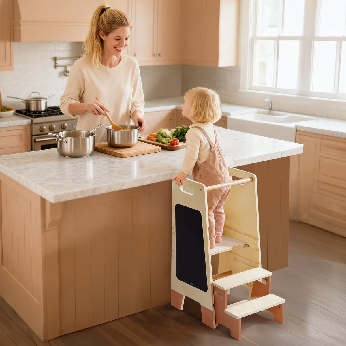 Smiling toddler in white outfit standing on ROBUD Toddler Standing Tower in a white kitchen with a chalkboard panel showing doodles