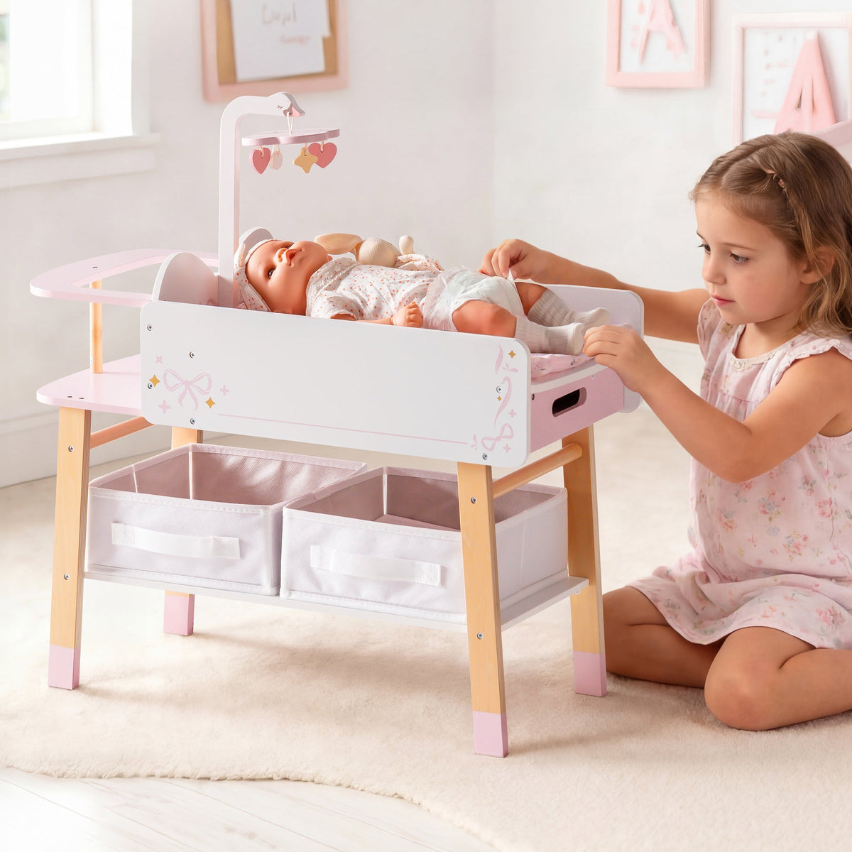 Young girl playing with ROBUD wooden doll changing table in white and pink with two pull-out storage baskets beneath.