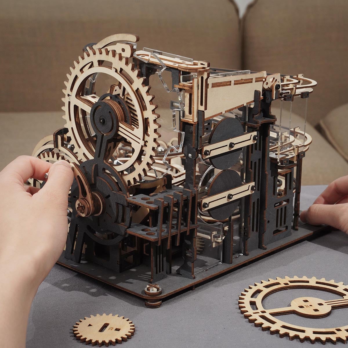 Hands pressing a large cog wheel into the ROKR Marble Night City tower with scattered wooden gears on a grey surface nearby