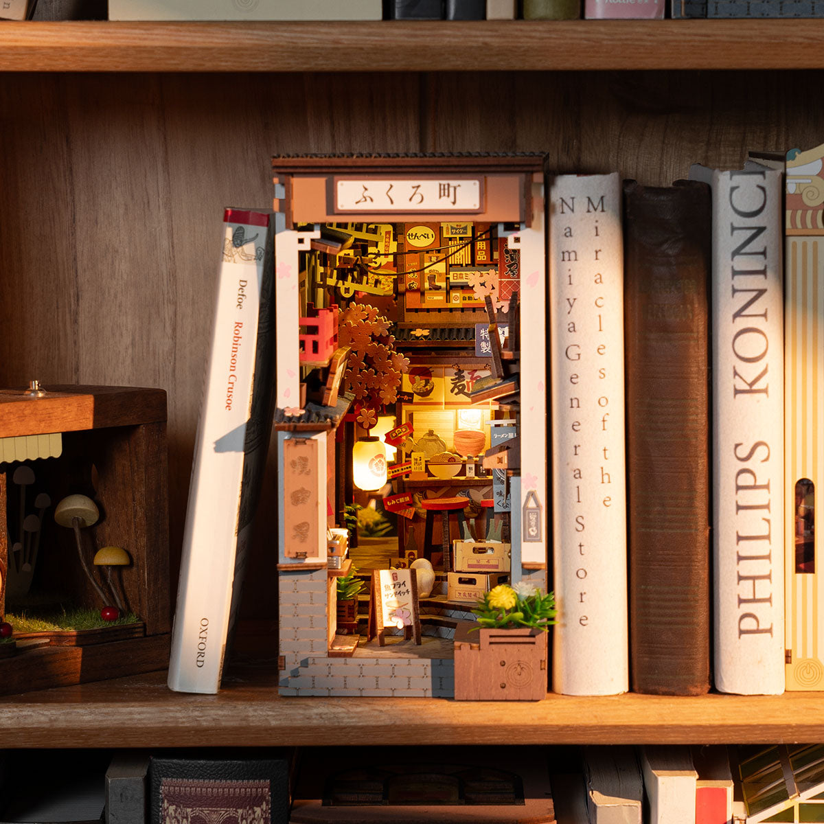 Rolife Sakura Wine Alley book nook TGB11 lifestyle scene on a desktop surrounded by books showing warm glowing Japanese street scene with cherry blossom and miniature figures