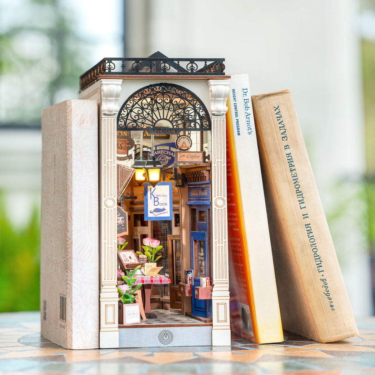 Rolife Arcade Dating Book Nook nestled between real books on a shelf, showing arched storefront facade with potted plants