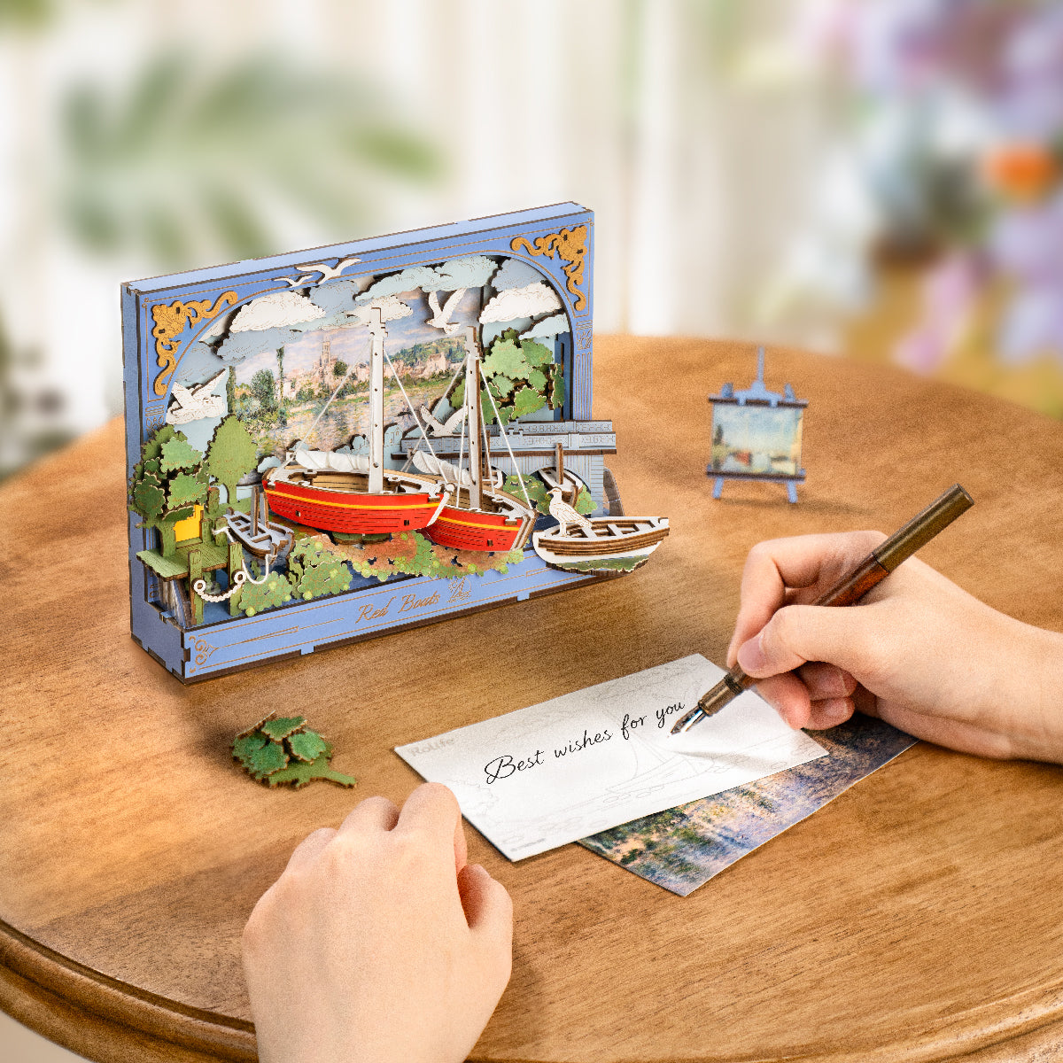 Hand writing a postcard beside the assembled Rolife Red Boats 3D art frame on a wooden desk with leaves