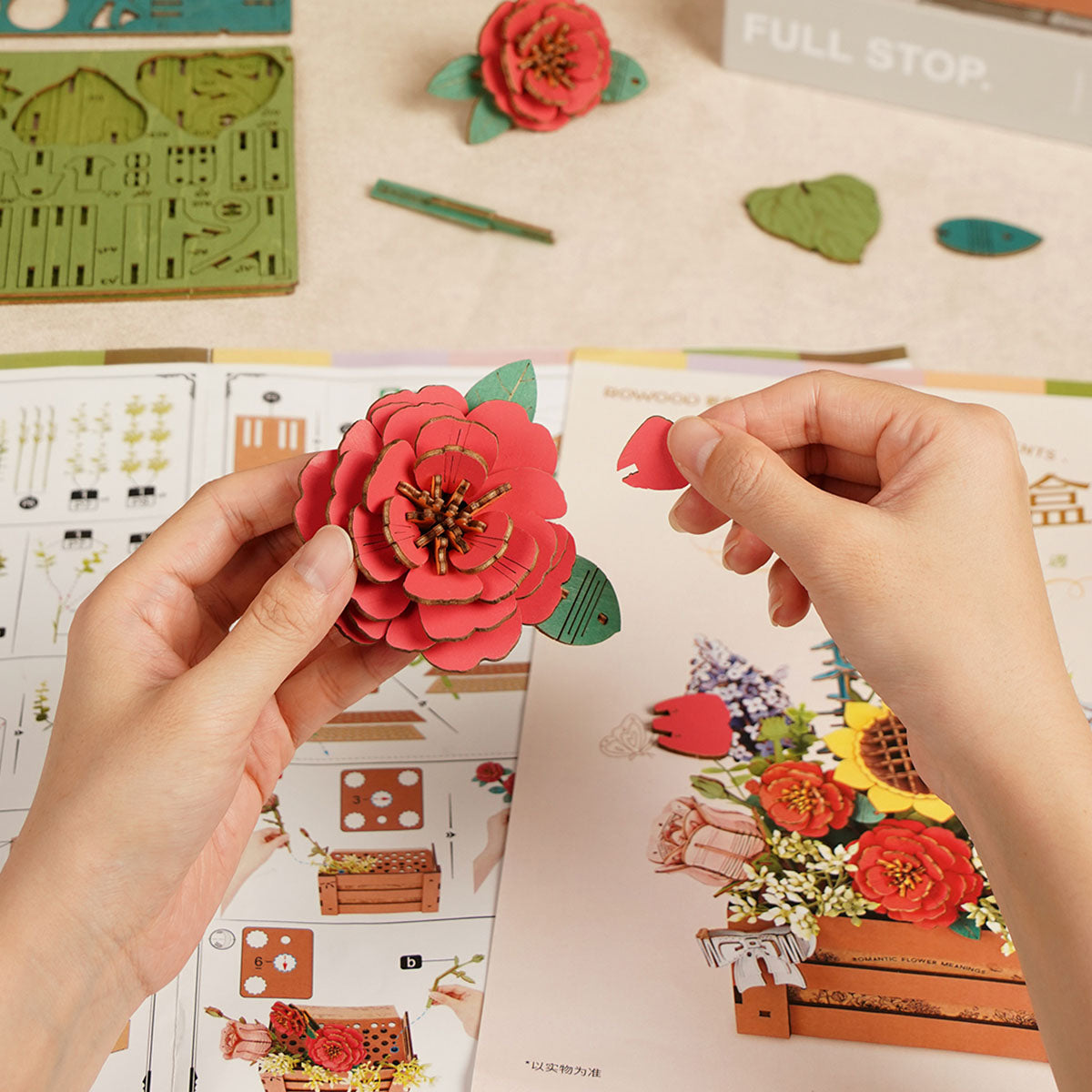 Hands assembling a Rowood wooden flower bloom, red camellia petals being clicked together above instruction sheet