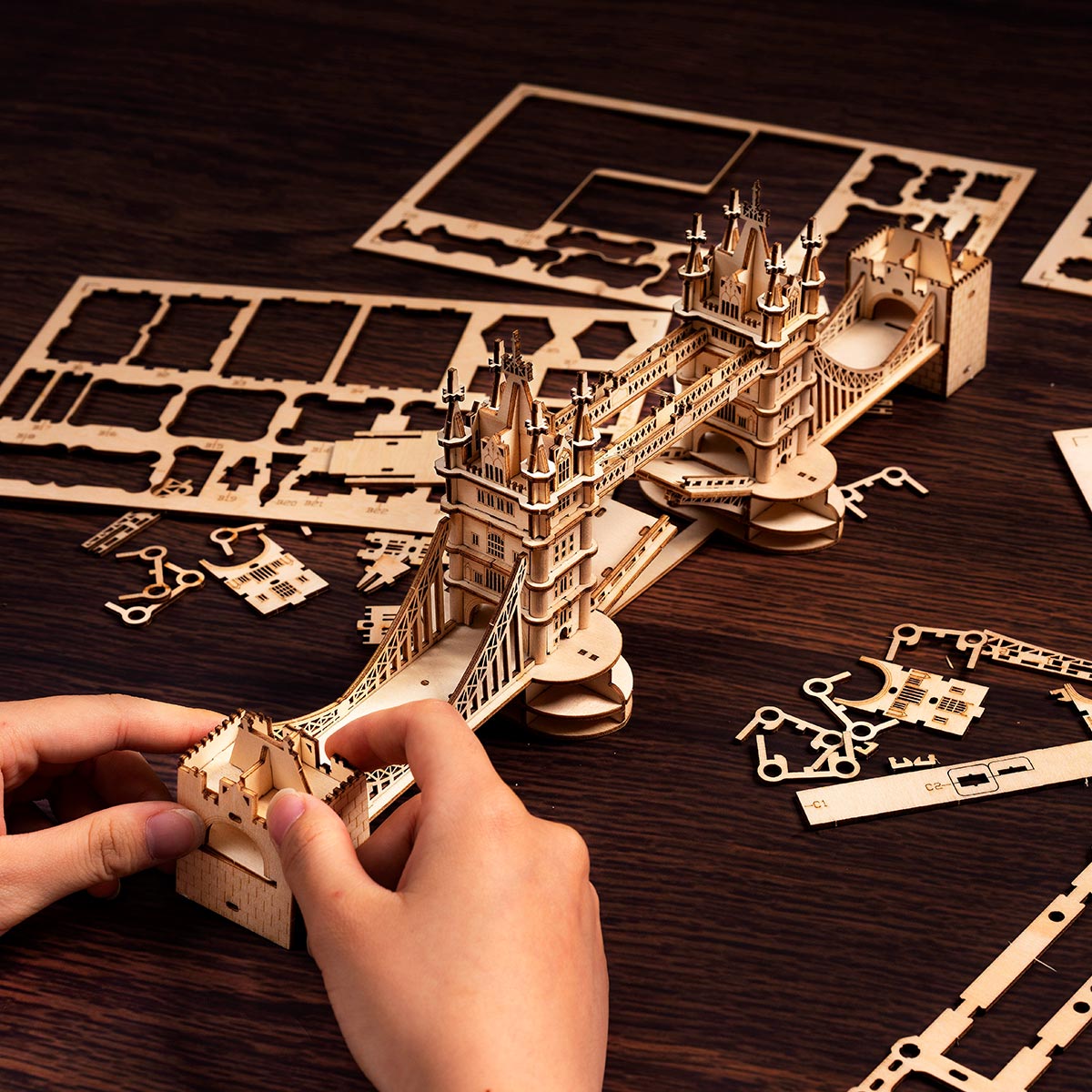 Hands assembling the Rolife Tower Bridge wooden puzzle with laser-cut plywood sheets spread across a dark walnut table