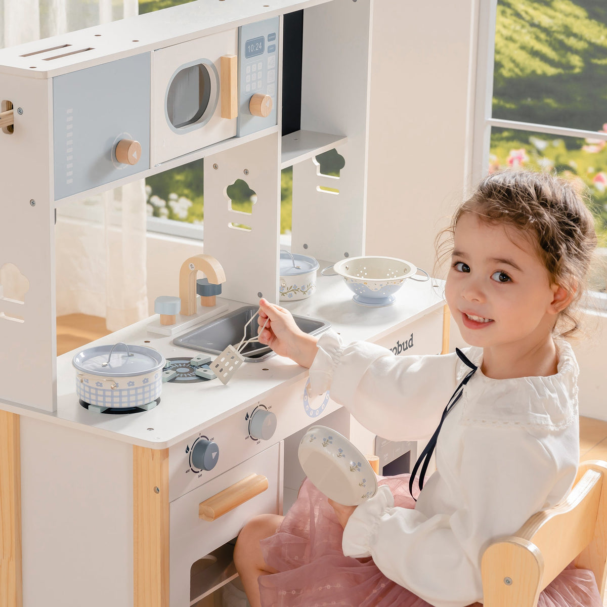 Smiling girl plays at the ROBUD white wooden kitchen, arranging a rice cooker and metal bowls on the countertop.