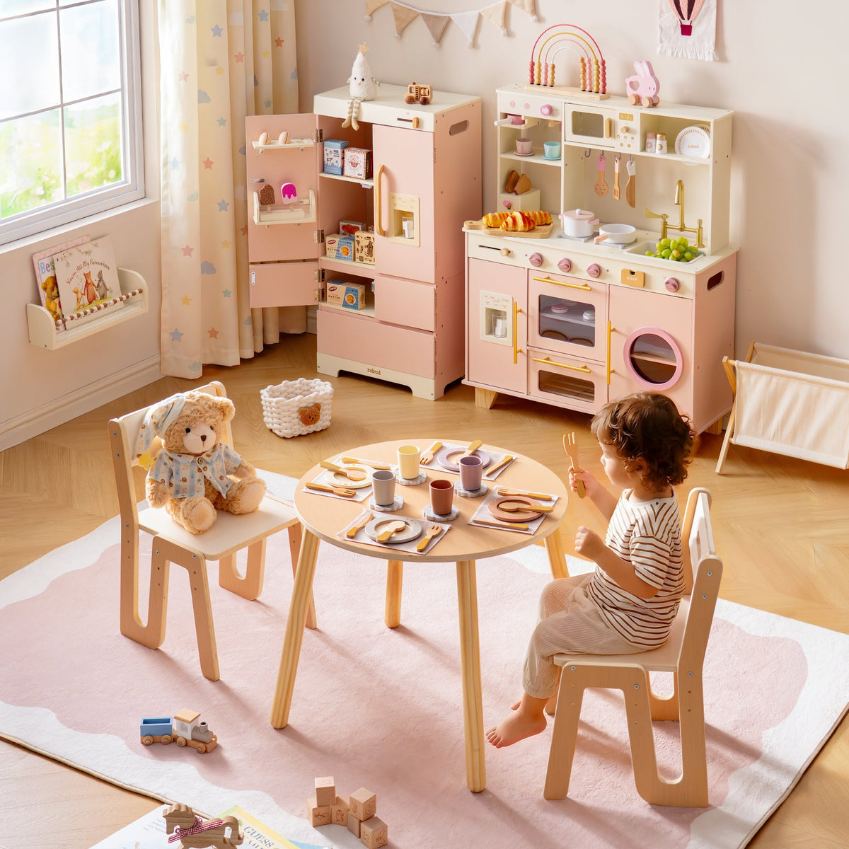 Child setting wooden dishes on a round ROBUD Glowwood table with the pink kitchen unit and toy food visible in the background.