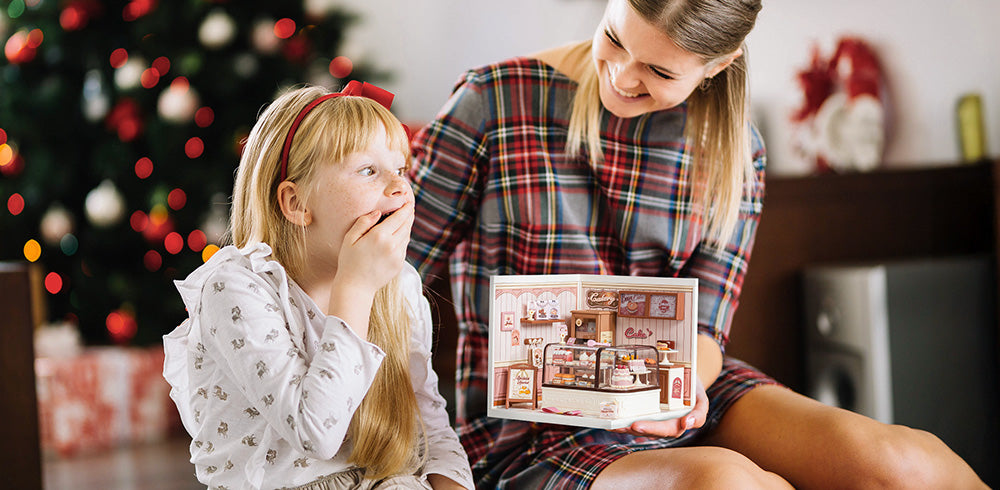 Mother presenting a Robotime miniature bakery house kit to a wide-eyed girl by a glowing Christmas tree, Robotime for kids desktop promotional banner