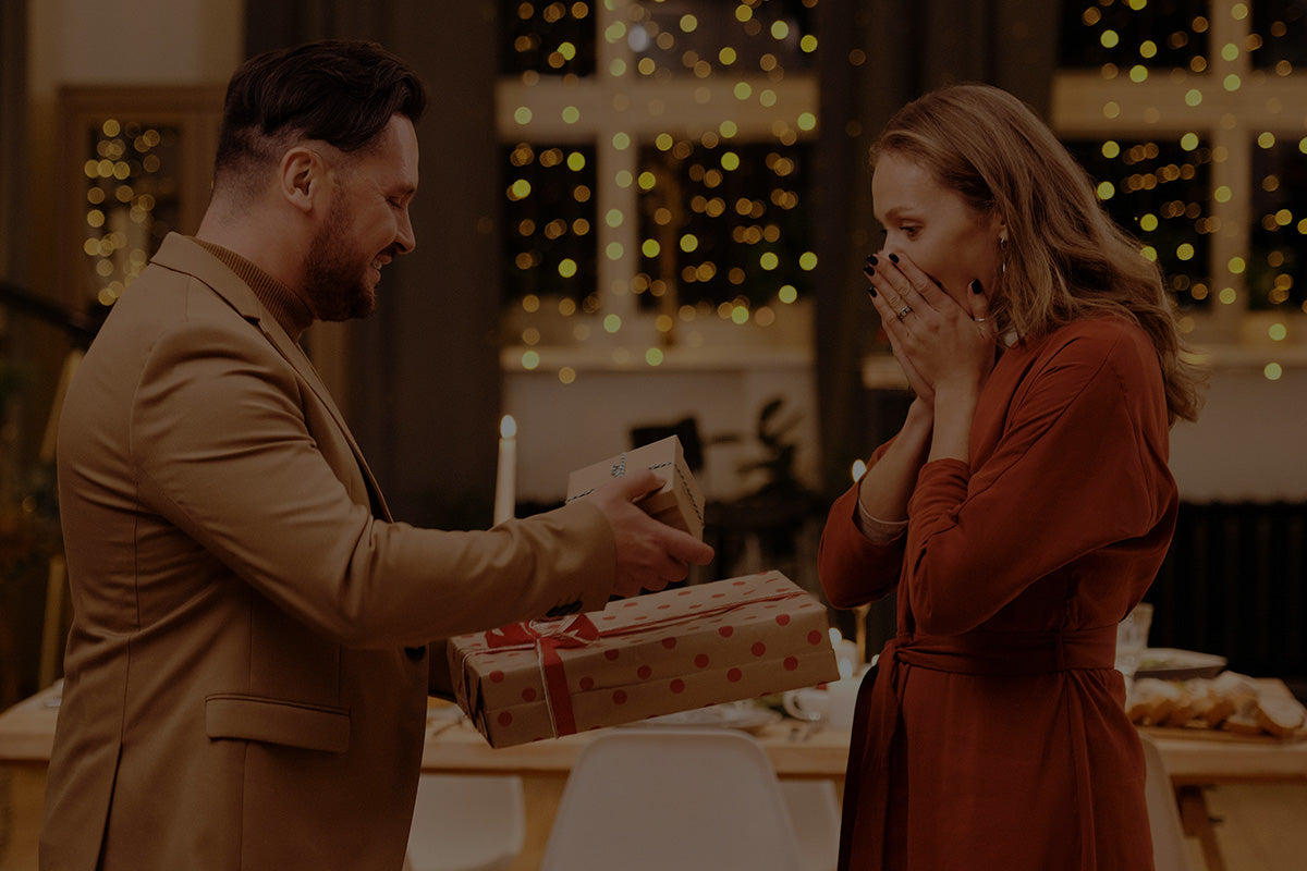 Robotime Online Gifts for Your Lover, man presenting gift boxes to a surprised woman in red at a candlelit dinner