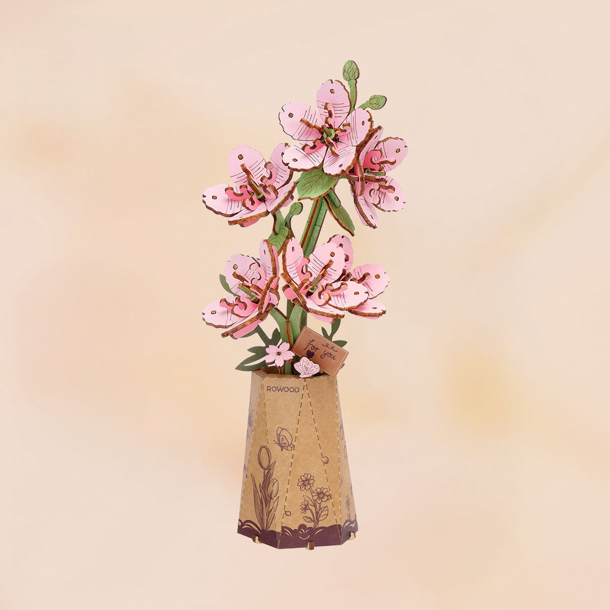 Rowood DIY-Holzblumenstrauß TW101: fertiges Kirschblüten-Modell mit üppigem rosa Sakura-Zweig-Arrangement in einem Kraftpapier-Topf vor pfirsichfarbenem Hintergrund.