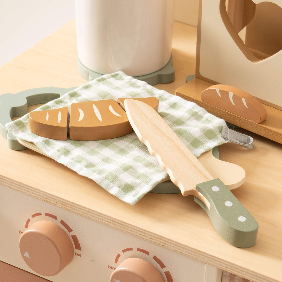 ROBUD wooden toy knife set with sage green handle resting on a checkered napkin beside sliced baguette on a birchwood toy kitchen counter
