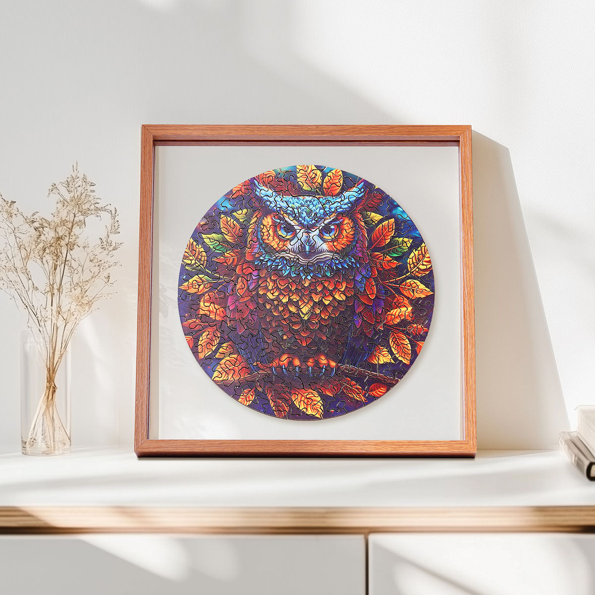 Rowood Moonlight Owl circular wooden jigsaw puzzle in a shadow box frame on a shelf beside a glass vase with dried stems and a candle