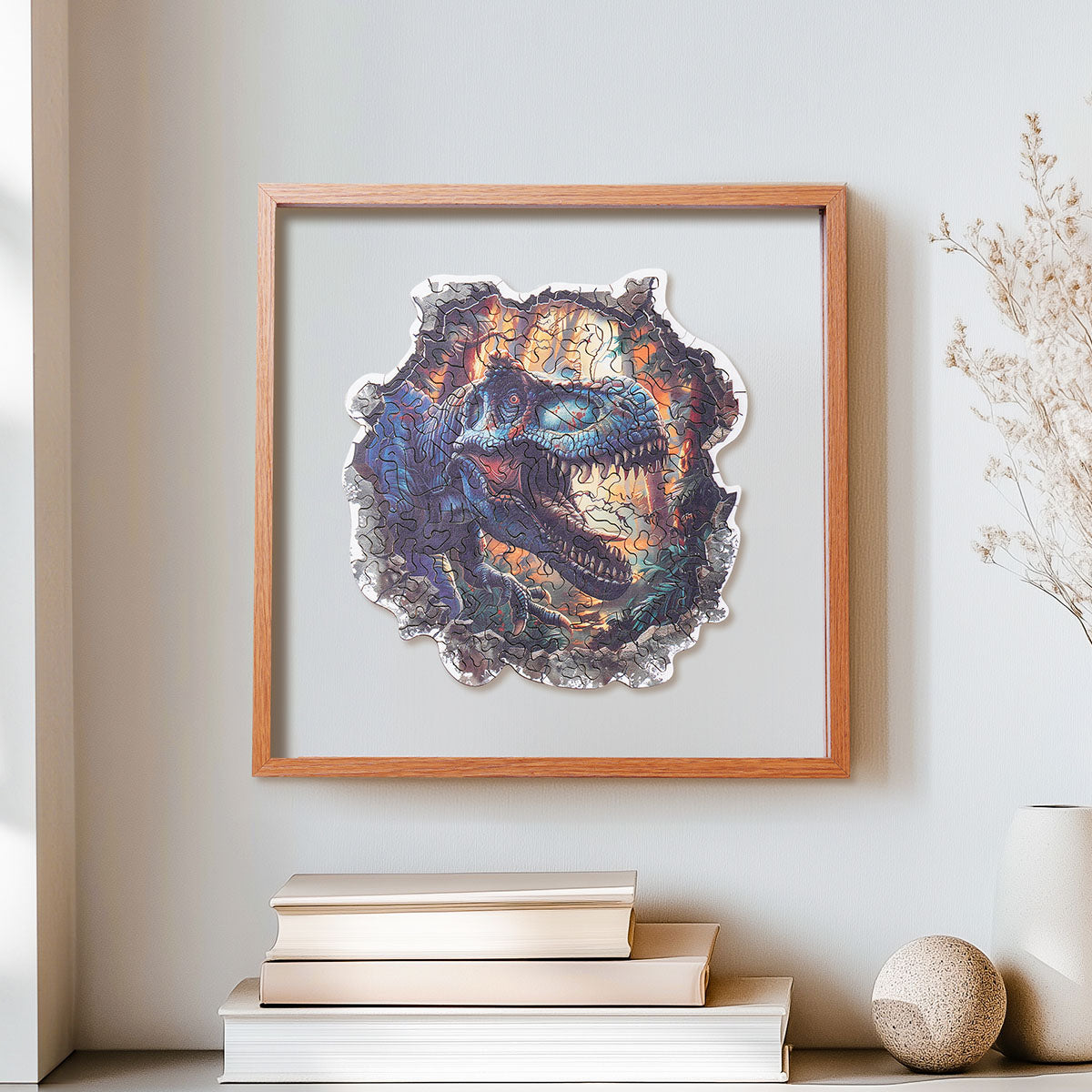 Rowood Wall Breaking T-Rex wooden jigsaw puzzle in a shadow box frame mounted on a wall above a stack of books and a round stone ornament