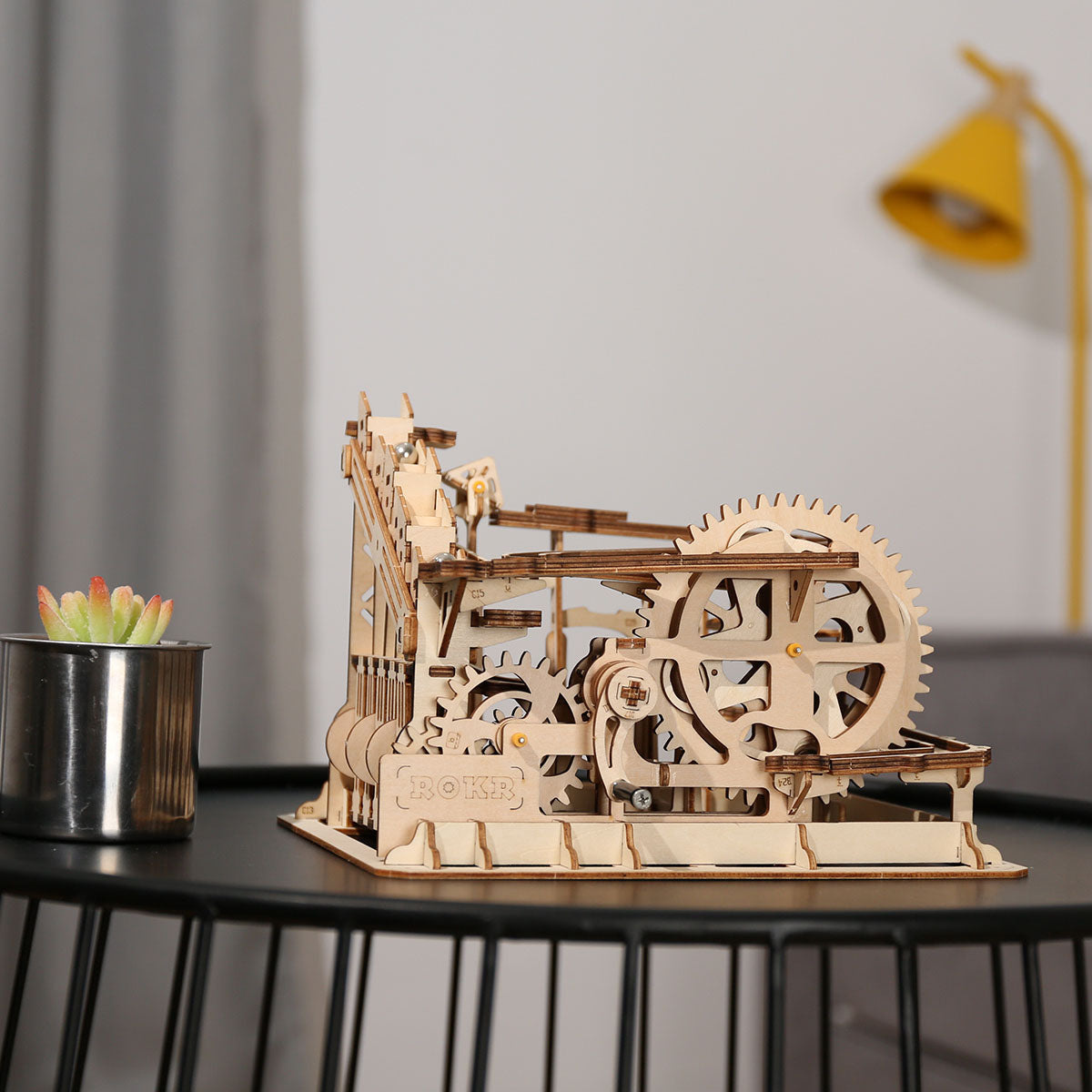 ROKR wooden marble run with large drive gear and tiered ramp tracks on a black table beside a yellow desk lamp