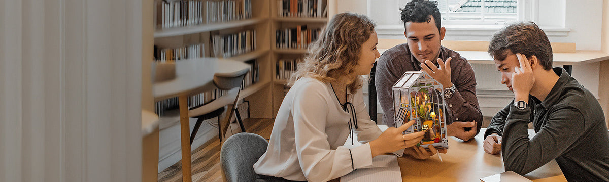 Wide-angle lifestyle scene of three people at a library table admiring a small illuminated Robotime miniature house with bookshelves behind them