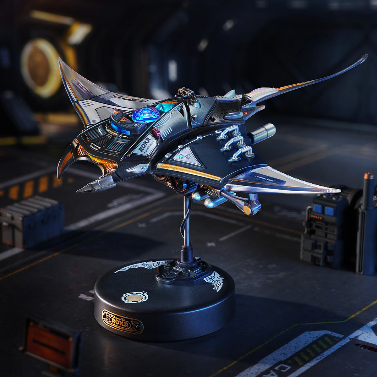 ROKR Manta Ray MI06 completed dark metallic model on its round branded stand displayed in a sci-fi hangar environment with blue and gold wing detail