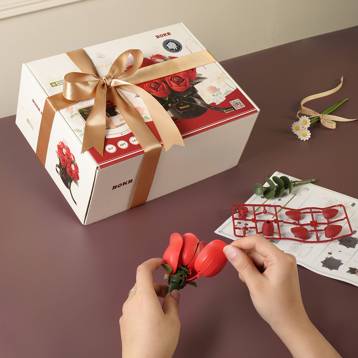 ROKR Red Rose Bouquet DIY kit AF02J gift box with gold satin bow beside hands assembling a red plastic rose bud with green stem runner parts and an instruction sheet