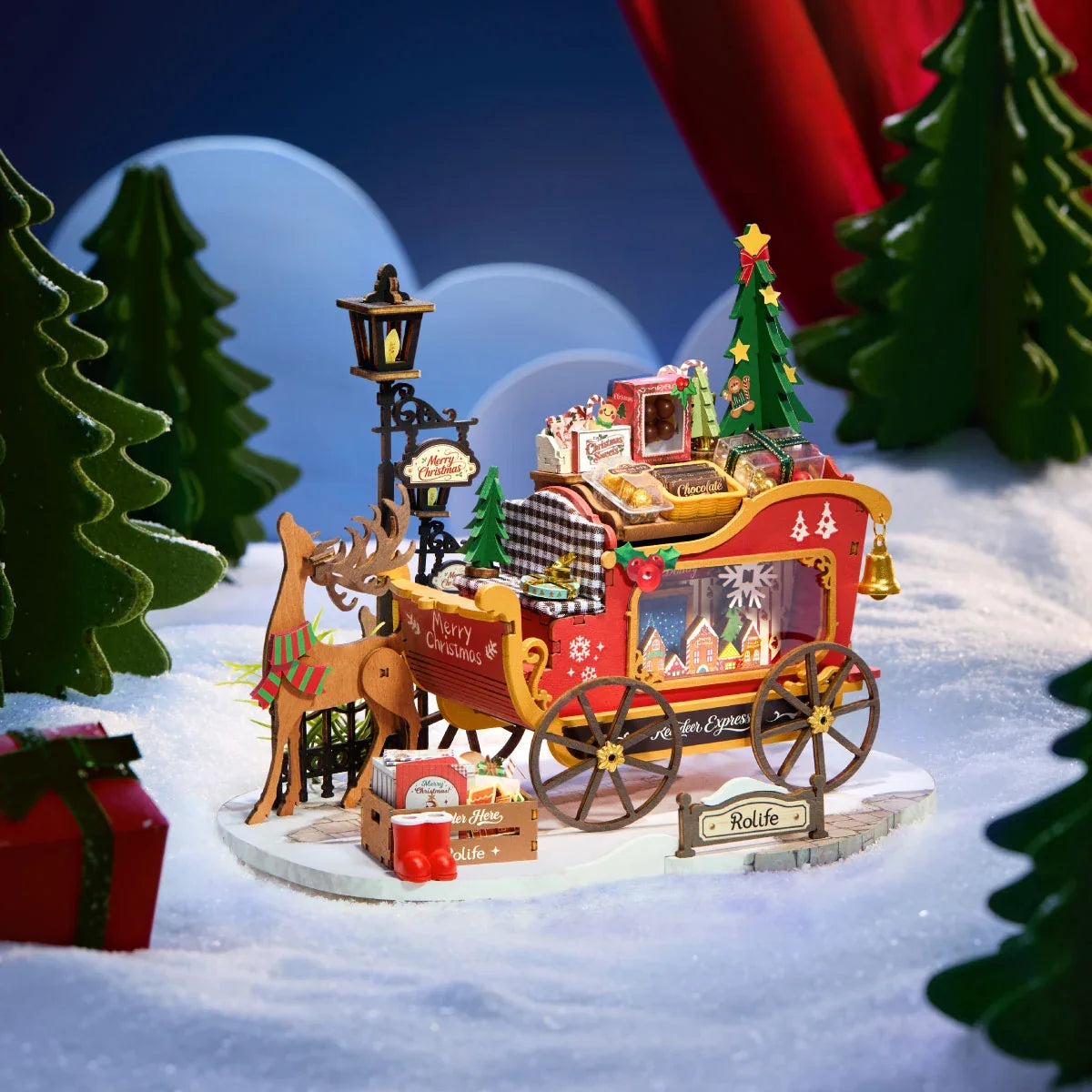 Rolife-Miniatur mit Holzrentier und rotem Schlitten auf verschneiter Basis, mit Laternenpfahl, Weihnachtsbaum, Geschenken und Süßigkeiten vor blauem Hintergrund mit grünen Bäumen.
