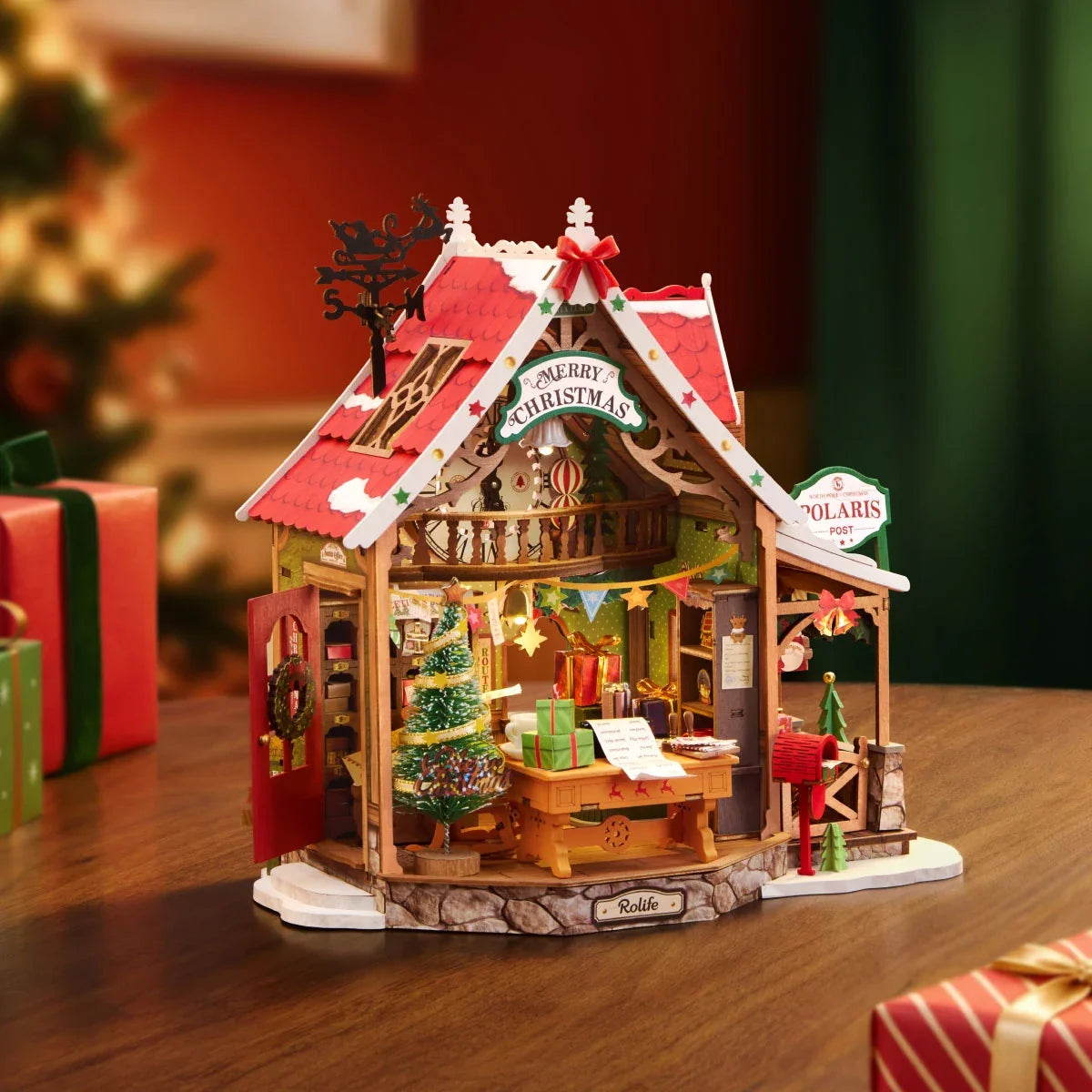 Rolife-Weihnachtspostamt im Miniaturformat auf einem Holztisch, mit offener Vorderseite, Weihnachtsmann am Schreibtisch, geschmücktem Baum, rotem Briefkasten und Polaris Post Schild sowie verpackten Geschenken daneben.