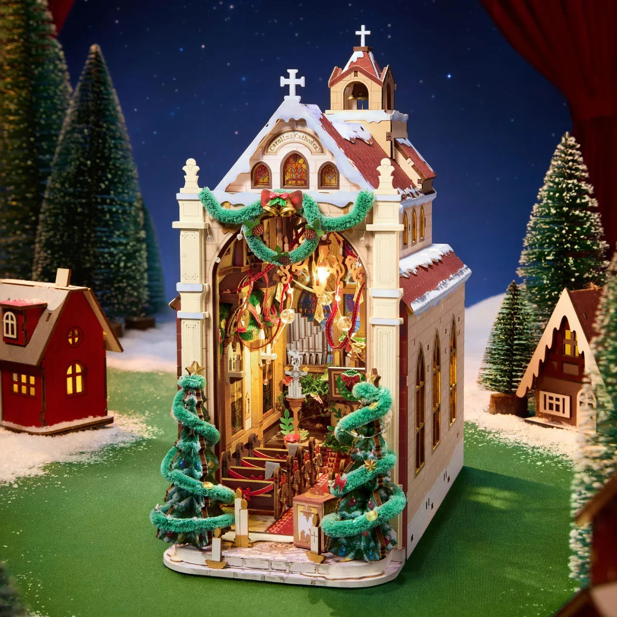 Rolife-Weihnachtskathedrale im Miniaturformat mit offener Vorderseite und beleuchtetem Innenraum, flankiert von geschmückten Bäumen auf einer verschneiten grünen Basis mit kleinen Häusern.