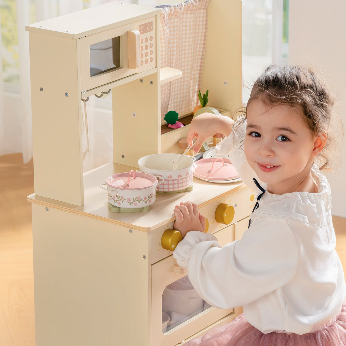 Smiling girl standing beside wooden ROBUD Tinplate Pretend Cooking Utensils pink play kitchen with sink, knobs and accessories in bright room