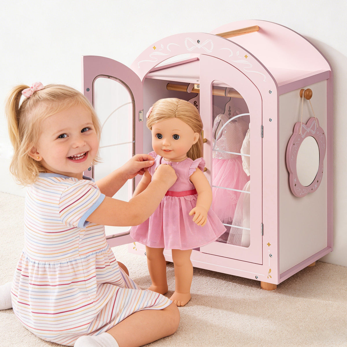 Young girl smiles as she places a doll into the ROBUD pink arched wardrobe in a pastel styled bedroom.