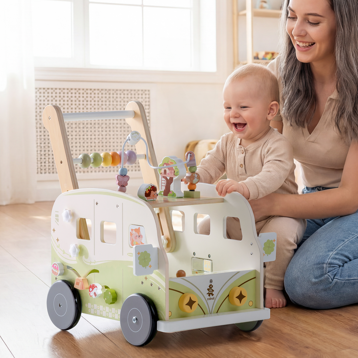 Toddler in a onesie takes steps pushing the ROBUD green wooden bus walker in a bright nursery with natural light.