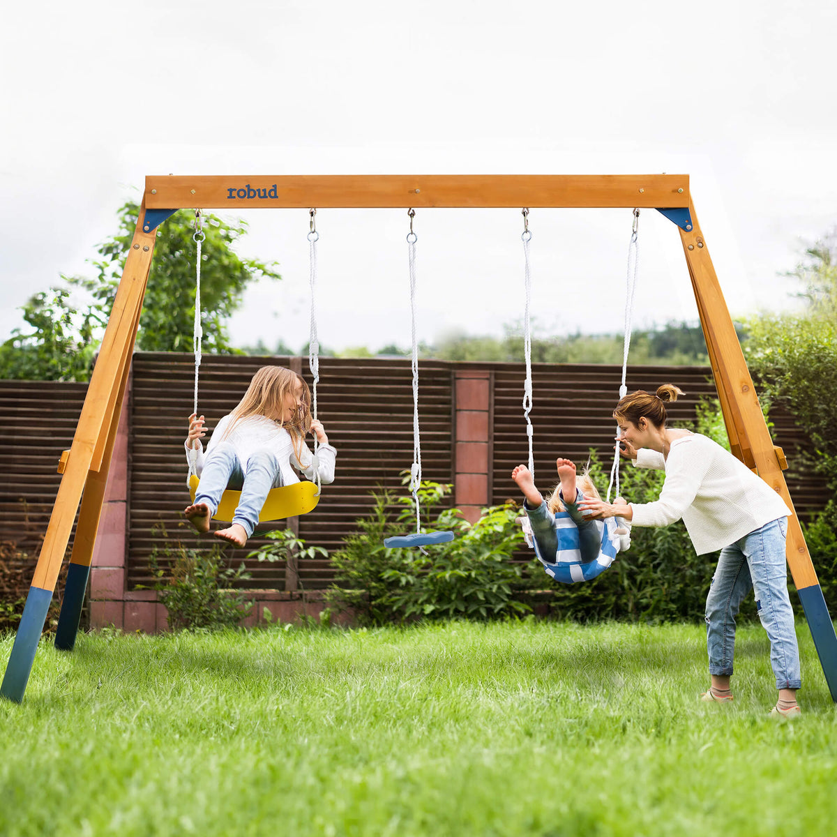 Mother pushing children on ROBUD honey-colored wood A-frame swing set in a backyard garden with yellow seat and blue baby bucket swing in use