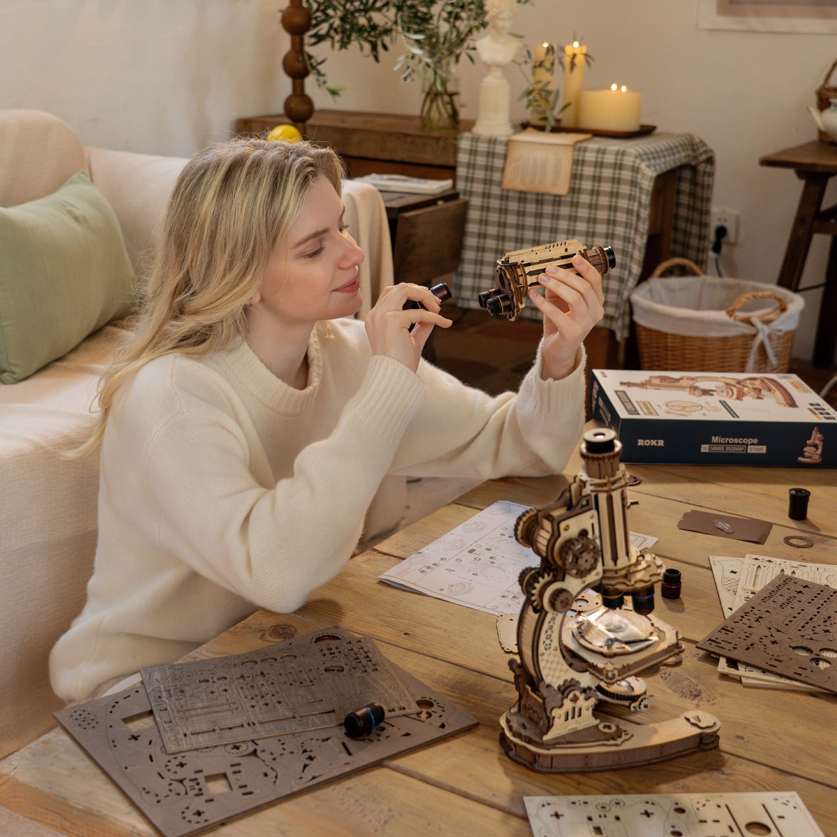 Woman in a cream sweater closely inspecting the ROKR wooden microscope model while seated at a cozy home workspace