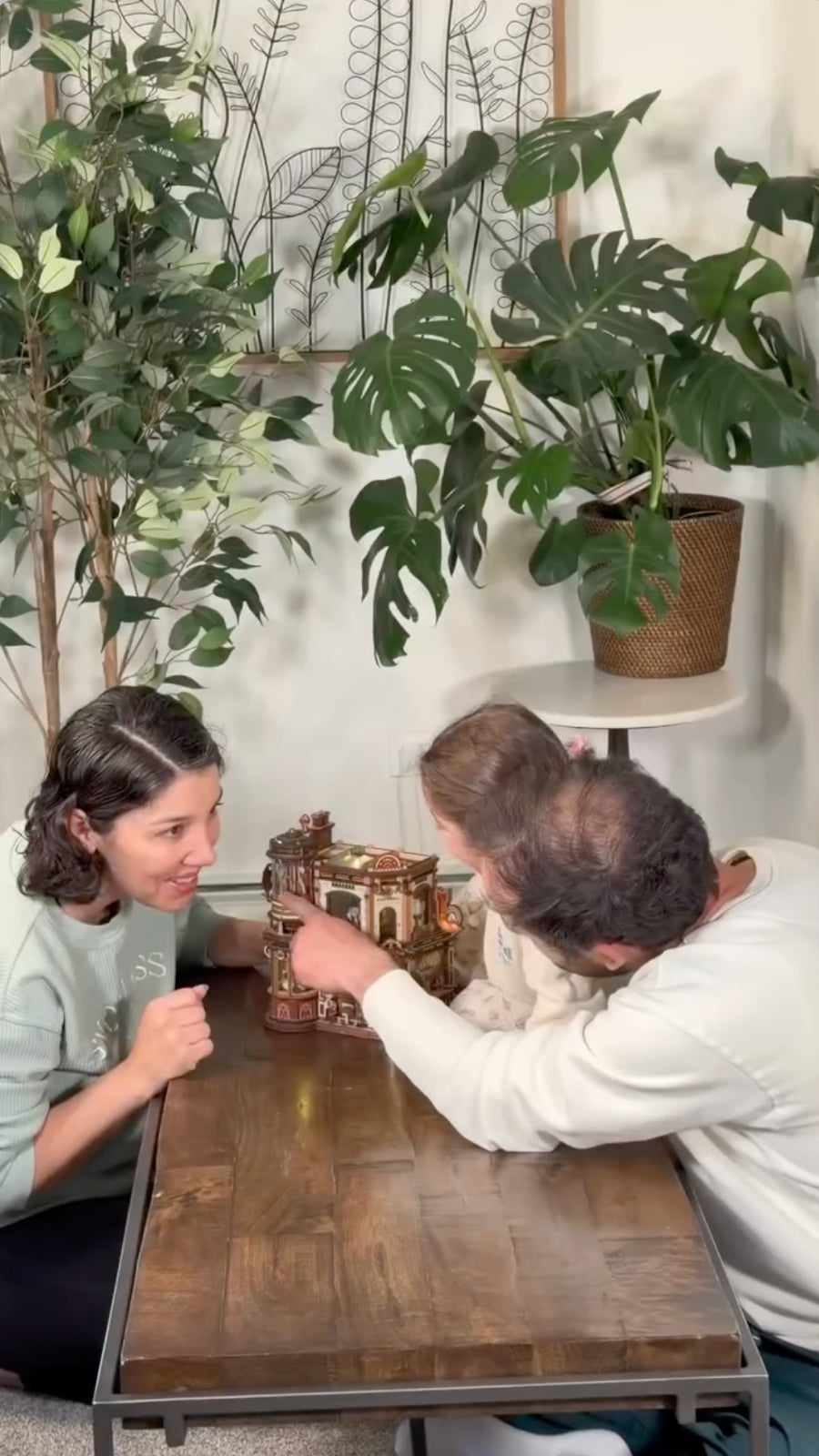 Two people assembling a wooden miniature together at a table surrounded by leafy plants