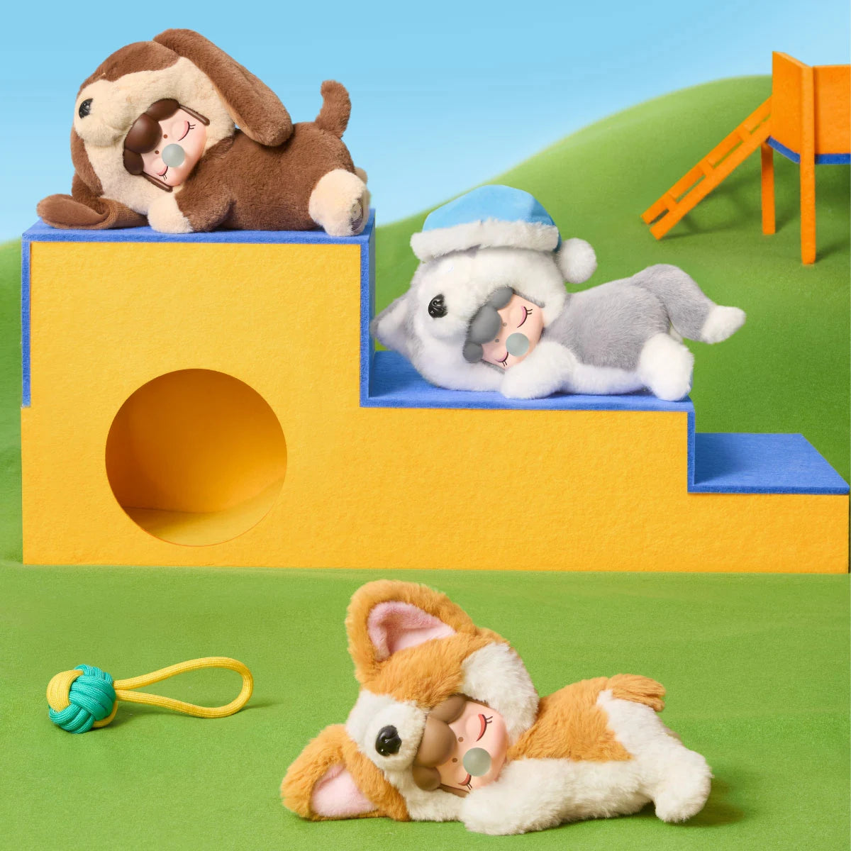 Drei Plüschfiguren in braunen, grauen und orangefarbenen Hundekostümen ruhen auf gelben und blauen Filzblöcken auf einer grünen Filzfläche mit einem Seilspielzeug.