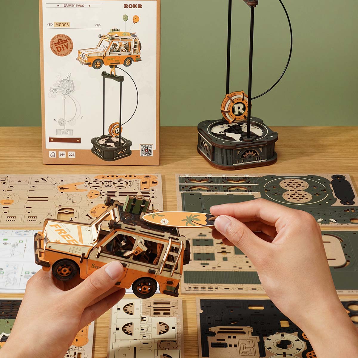 ROKR Road Tripper Gravity Swing MCD03 ROKR box packaging and partially assembled orange SUV body held by two hands, green part sheets spread on desk