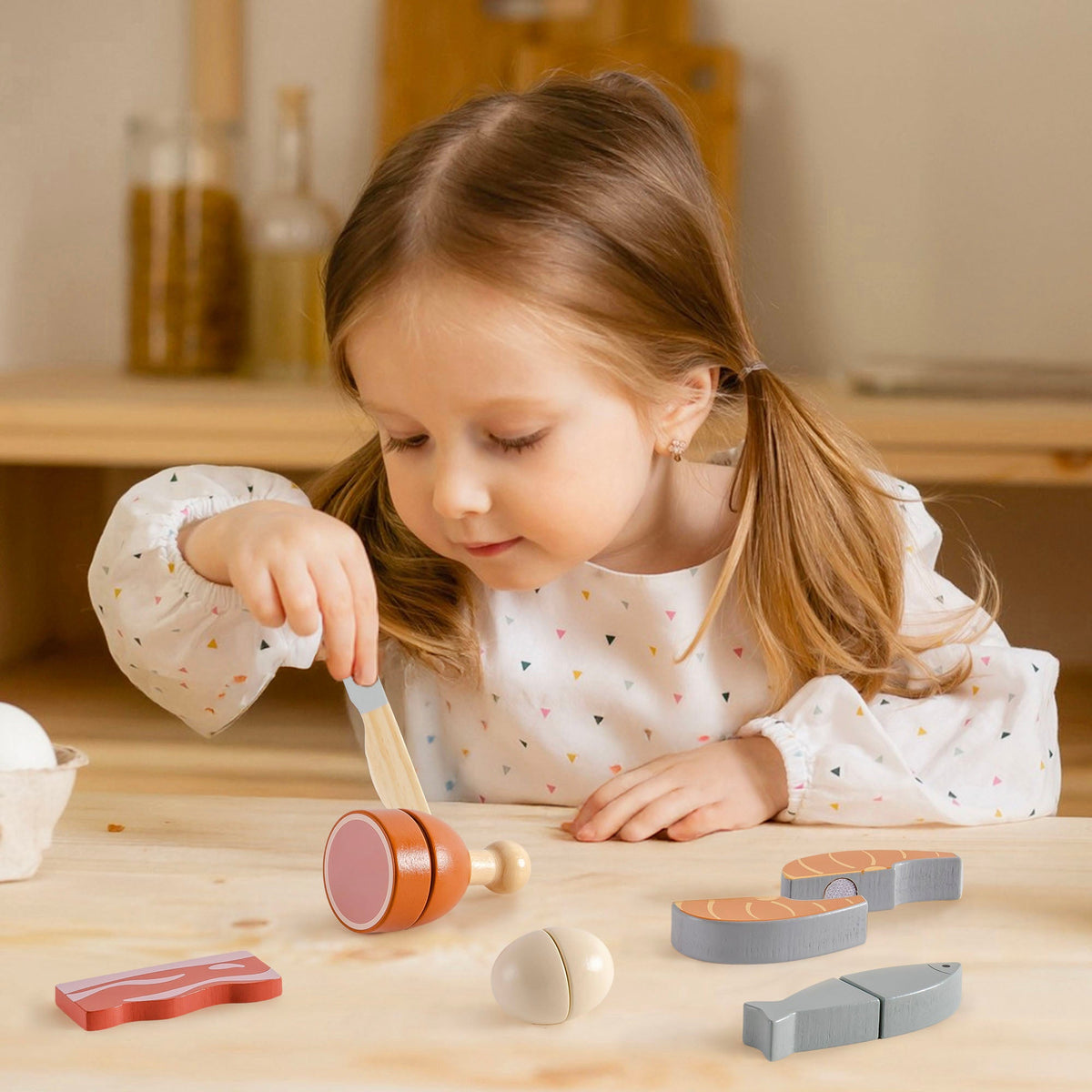 Young girl cutting a wooden chicken leg piece from the ROBUD meat playset on a wooden table with bacon strip, salmon halves, and grey fish visible nearby