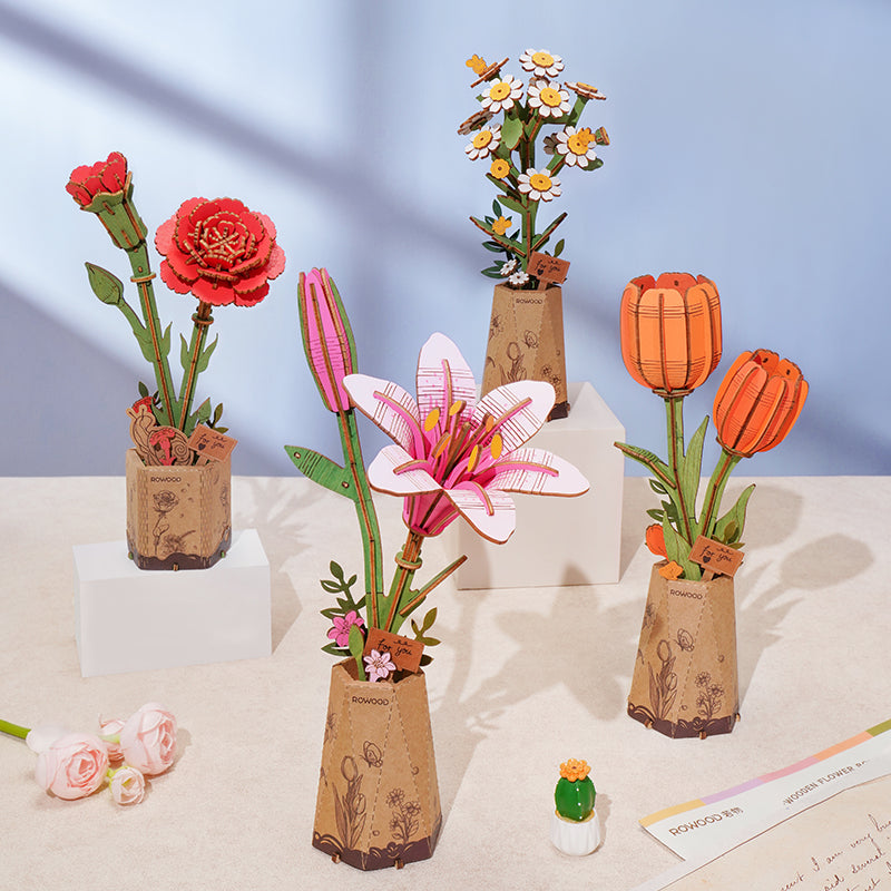 Four Rowood wooden flower puzzle models displayed on white pedestals showing a red carnation, a pink lily, white daisies, and an orange tulip in kraft paper pots