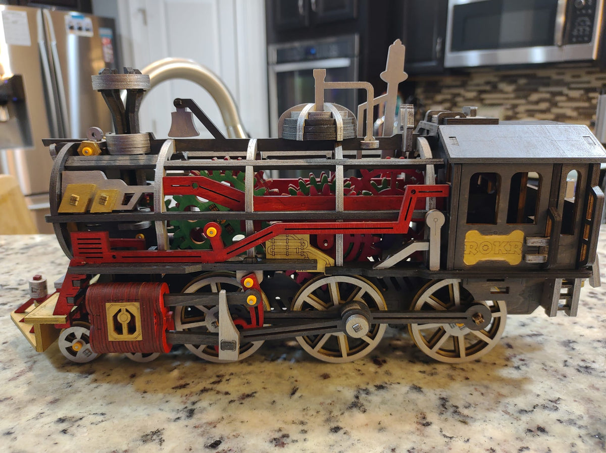 Robotime red steam locomotive model stands on a kitchen counter with brass details.