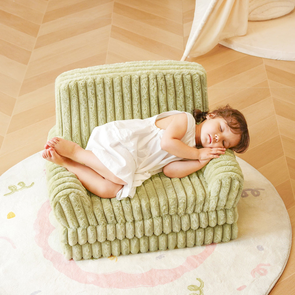 Toddler girl sleeping peacefully on a ROBUD green striped sofa in a soft white playroom with a round floor mat.