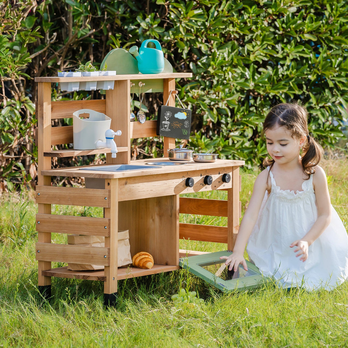 ROBUD Outdoor Mud Play Kitchen hero front view showing girl in white dress beside the full honey-toned wooden outdoor kitchen station in green garden
