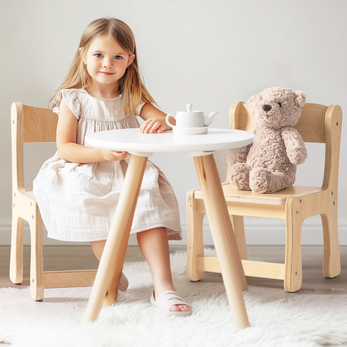 Young girl in a grey dress sitting at ROBUD white-top birch toddler table with a toy teapot and stuffed teddy bear on the adjacent chair