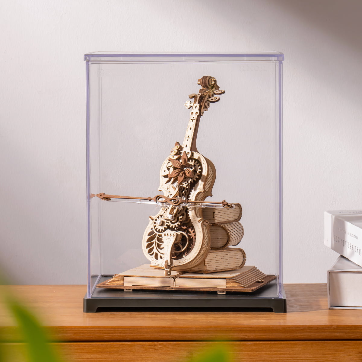 ROKR Magic Cello wooden music box in a clear display case placed on a wooden sideboard beside stacked books