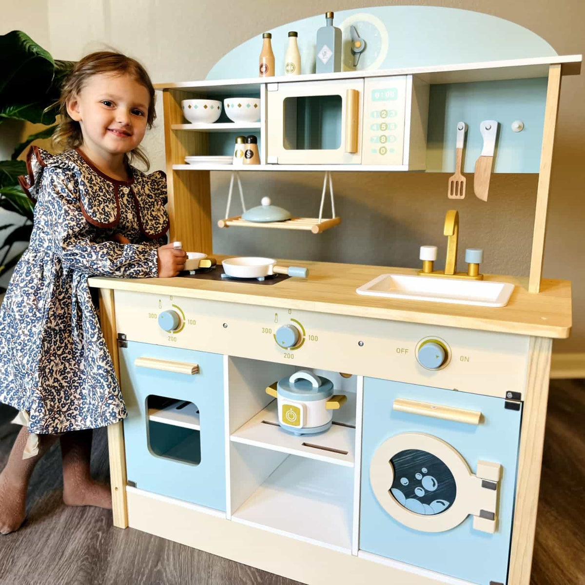 Young girl in a floral dress smiling beside a full-size ROBUD wooden kitchen play set in blue and natural wood with toy utensils, microwave, oven, sink, and washing machine in a home setting